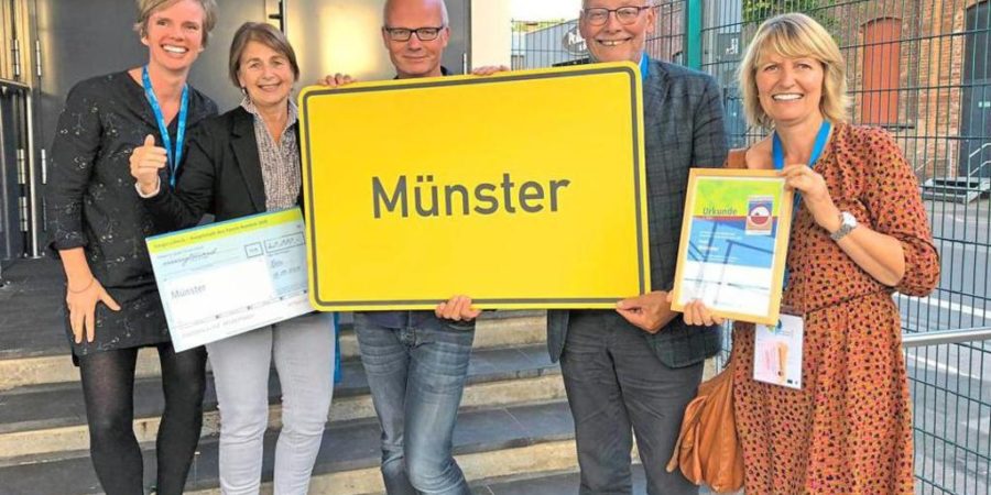 5 Personen der Fair Trade Steuerungsgruppe halten ein Gelbes Straßenschild mit dem Namen Münster hoch