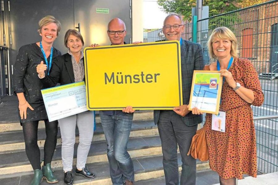 5 Personen der Fair Trade Steuerungsgruppe halten ein Gelbes Straßenschild mit dem Namen Münster hoch