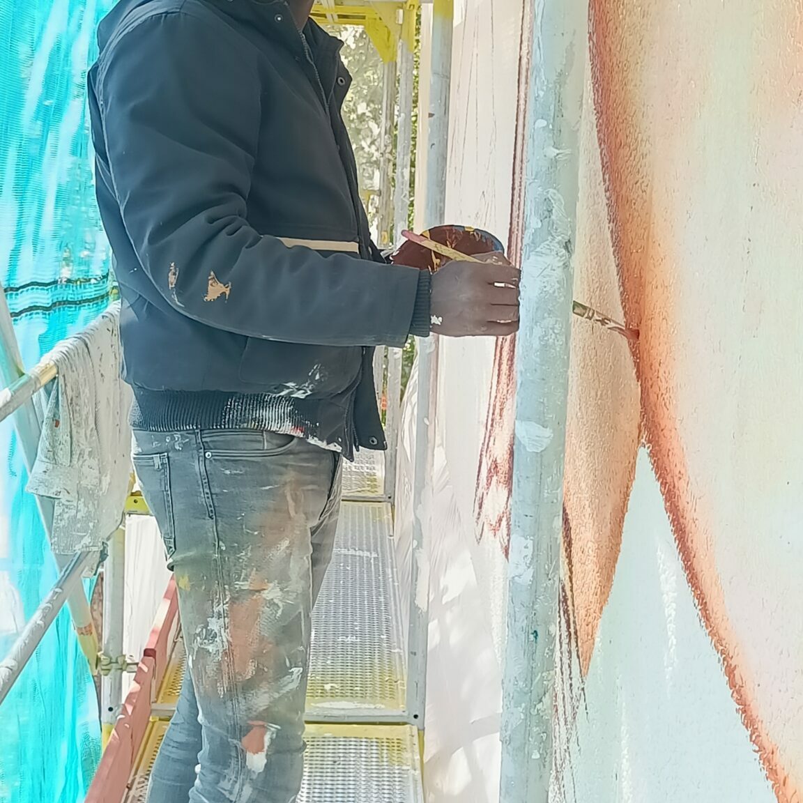 Der Künstler Daniel Kwaku Anetang steht auf einem Gerüst und hat einen Pinsel und eine Schüssel Farbe in der Hand.