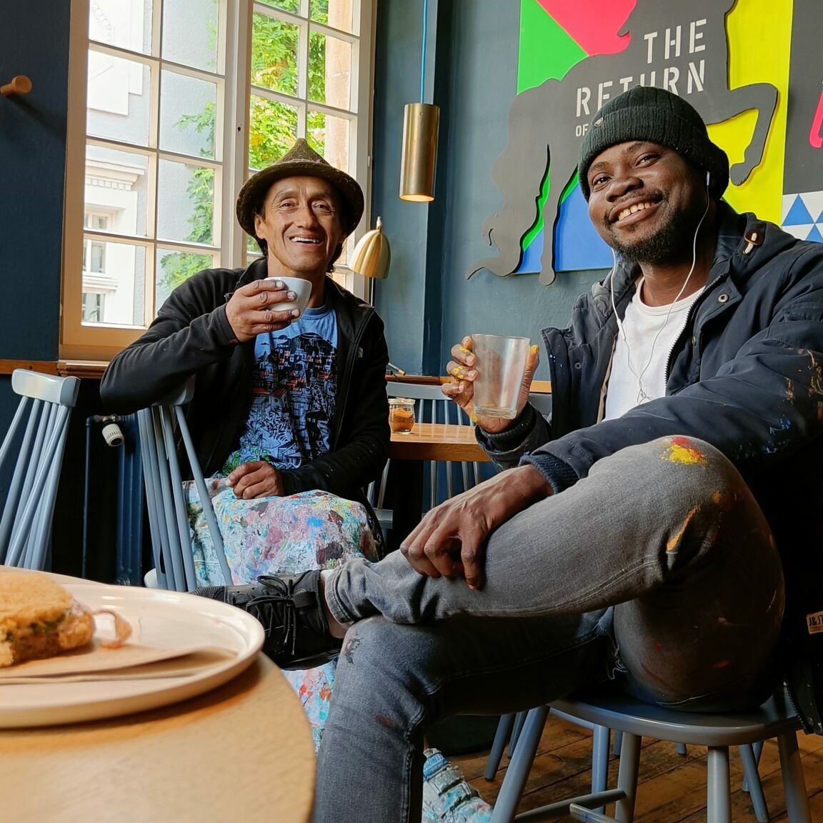 Die Künstler Daniel und Jorge sitzen in einem Café und trinken einen Kaffee zusammen. Sie lächeln beide in die Kamera