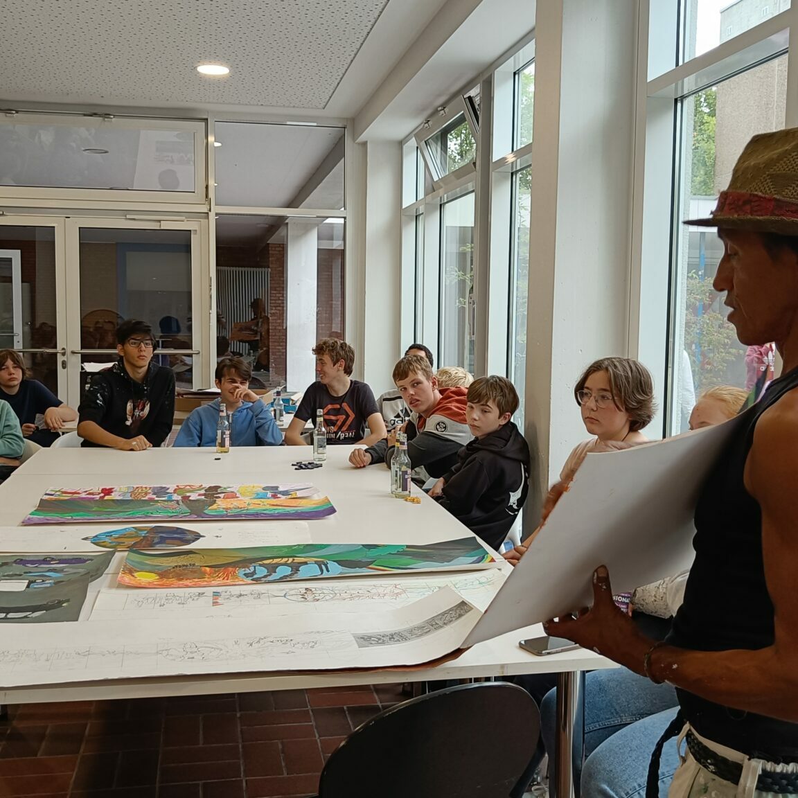 Bunte Skizzen liegen auf einem Tisch. Ein Mann mit Hut präsentiert sie Jugendlichen