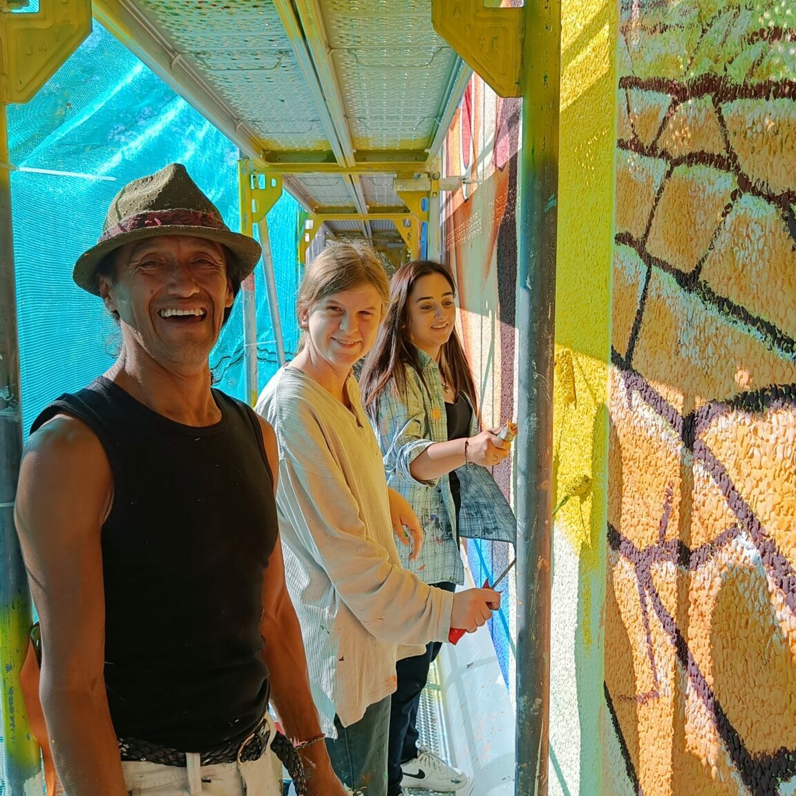 Ein Mann mit Hut steht auf einem Gerüst an einer Wand und lächelt in die Kamera. Hinter ihm stehen zwei jugendliche Mädchen mit Pinseln und lächeln.