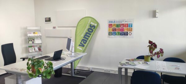 Zu sehen ist ein Büro mit Blumen, einer Beachflg mit dem Logo von Vamos und ein Plakat der SDGs.