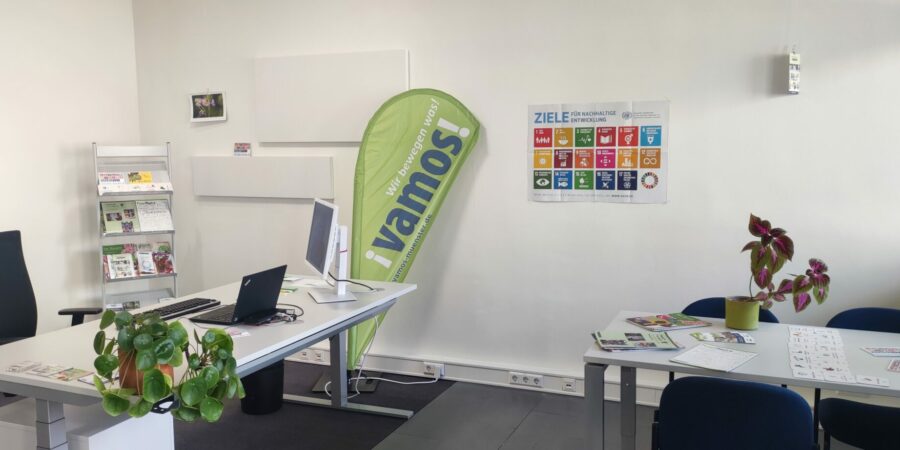 Zu sehen ist ein Büro mit Blumen, einer Beachflg mit dem Logo von Vamos und ein Plakat der SDGs.