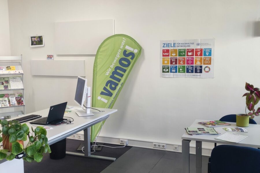 Zu sehen ist ein Büro mit Blumen, einer Beachflg mit dem Logo von Vamos und ein Plakat der SDGs.