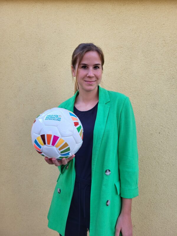 Frau steht in einem grünen Blazer vor einer Wand und lächelt in die Kamera, in der Hand trägt sie einen Fußball mit der Aufschrift "Ziele für Nachhaltige Entwicklung"